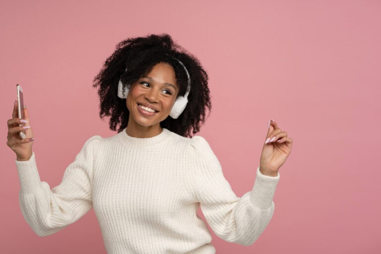 Happy dark skinned millennial woman with headphones enjoying listening to the artist's new album, dances, makes movements to music, holding mobile phone, isolated on studio pink background. Copy space